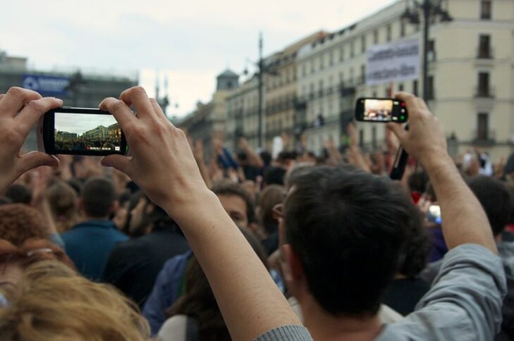 Zu sehen ist eine Ansammlung vieler Menschen. Zwei Personen im Vordergrund sind von hinten abgebildet und halten je ein Smartphone in die Höhe, mit dem sie das Geschehen aufzeichnen.