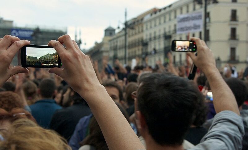 Zu sehen ist eine Ansammlung vieler Menschen. Zwei Personen im Vordergrund sind von hinten abgebildet und halten je ein Smartphone in die Höhe, mit dem sie das Geschehen aufzeichnen.
