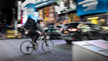 Ein Mensch fährt auf einem Fahrrad im Straßenverkehr und hat eine Box auf dem Rücken, die typischerweise im Lieferdienst benutzt wird.
