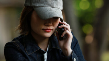 Eine Frau hält ein Smartphone an ihr Ohr. Sie trägt eine Kappe, die einen Schatten auf den oberen Teil des Gesichts wirft.