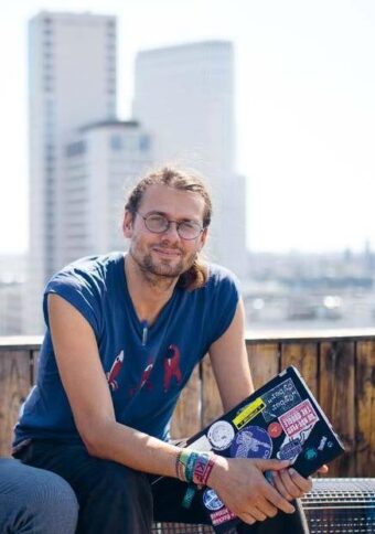 Auf dem Bild ist der Interviewte, Rainer Rehak, zu sehen. Rainer sitzt auf einer Bank und hält sein mit Aufklebern beklebtes Notebook in der Hand. Im Hintergrund sind Hochhäuser zu sehen.