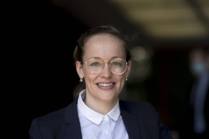 Eine junge Frau mit Brille lächelt in die Kamera. Sie trägt ein dunkles Jacket und ein weißes Hemd.