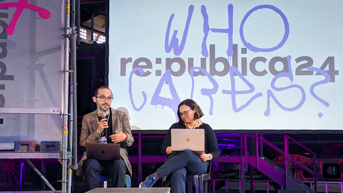 Fabio Chiusi und Antonella Napolitano sitzen auf einer Bühne. Hinter ihnen ist auf einer Leinwand das Logo der Republica 2024 abgebildet, überlagert mit einem Schriftzug des Mottos "Who Cares?". Chiusi, links im Bild, spricht in ein Mikrophon und gestikuliert. Napolitano hat ein aufgeklapptes Macbook auf dem Schoß.