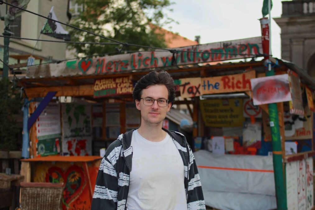 Ein Mann Mitte 20 mit weißem T-Shirt, Brille und schwarzen Haaren steht vor einem aus Holz selbstgebauten Infostand des Klima-Camps Augsburg. Auf dem Klima-Camp stehen Botschaften wie "Tax the Rich" und "Herzlich Willkommen".