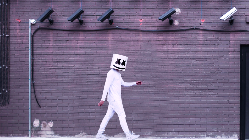 Eine weiß gekleidete Person mit einem weißen Eimer auf dem Kopf läuft unter Überwachungskameras an einer Mauer entlang