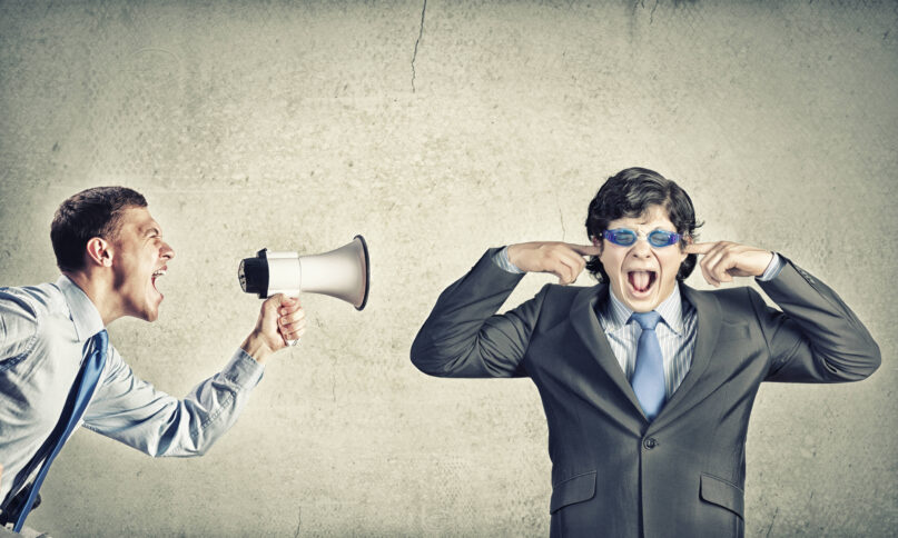 Ein Mann im Hemd schreit durch ein Megafon einen zweiten Mann im Anzug an, der eine Schwimmbrille aufhat und sich die Ohren zuhält.