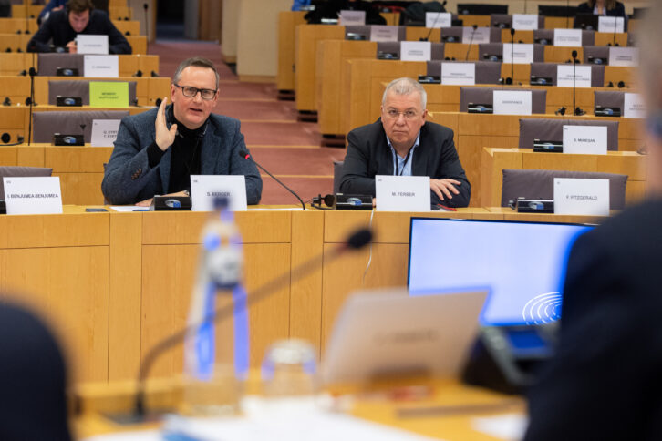 Stefan Berger und Markus Ferber sitzen in einem Ausschusssaal des EU-Parlaments. Berger argumentiert, Ferber hört zu.