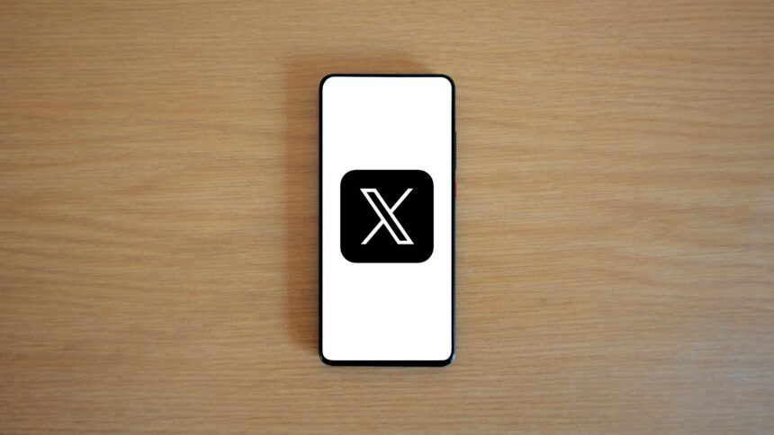 Ein Handy liegt auf einem Holztisch. Es zeigt das Logo von X, früher Twitter.