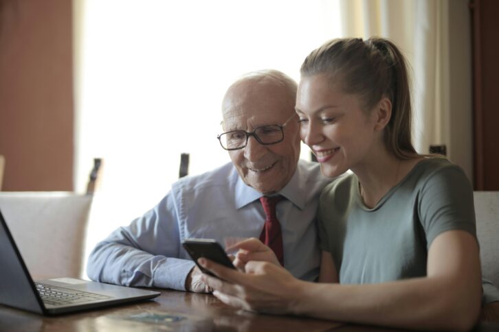 A young woman shows something to an old man on a phone. They have a laptop in front of them.