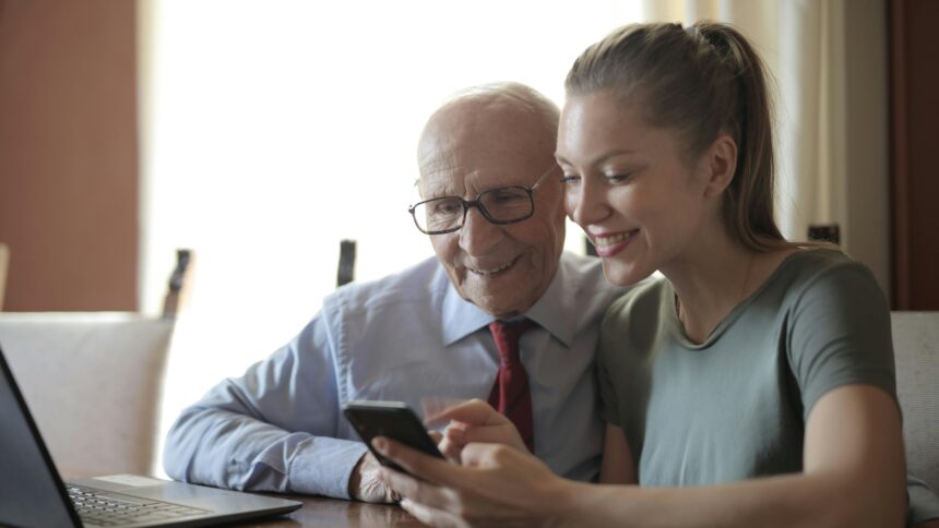 A young woman shows something to an old man on a phone. They have a laptop in front of them.