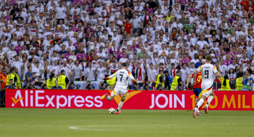 Ein Fußballspieler auf dem Feld, kurz vor dem Abschuss. Im Hintergrund ein AliExpress-Werbebanner und Tribünen voller Zuschauer.