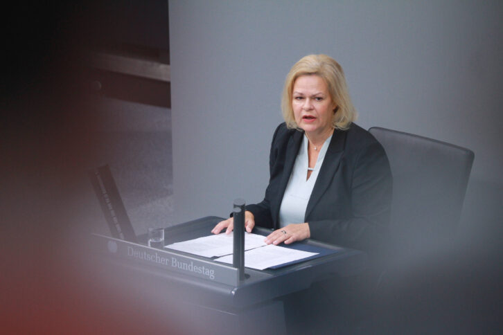 Person mit blonden Haaren im schwarzen Blazer an Pult