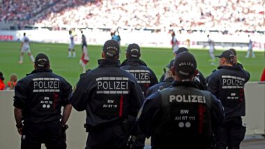 Mehrere Polizisten stehen mit dem Rücken zum Bild, im Hintergrund ein Fußballspiel im Stadion