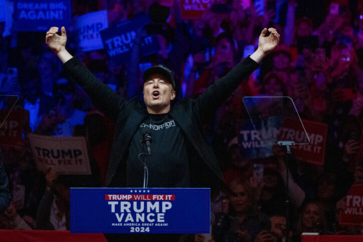 Mann mit schwarzer Kappe steht an Pult und reißt die Arme hoch