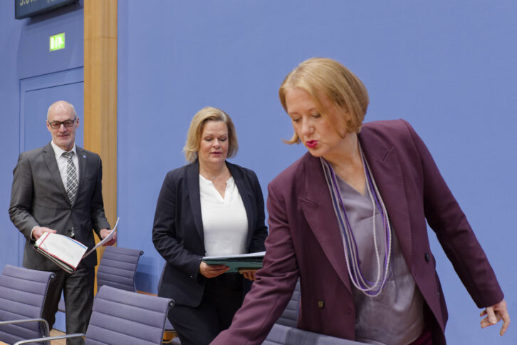 Drei Menschen vor blauem Hintergrund, vorne eine blonde im lila Blazer