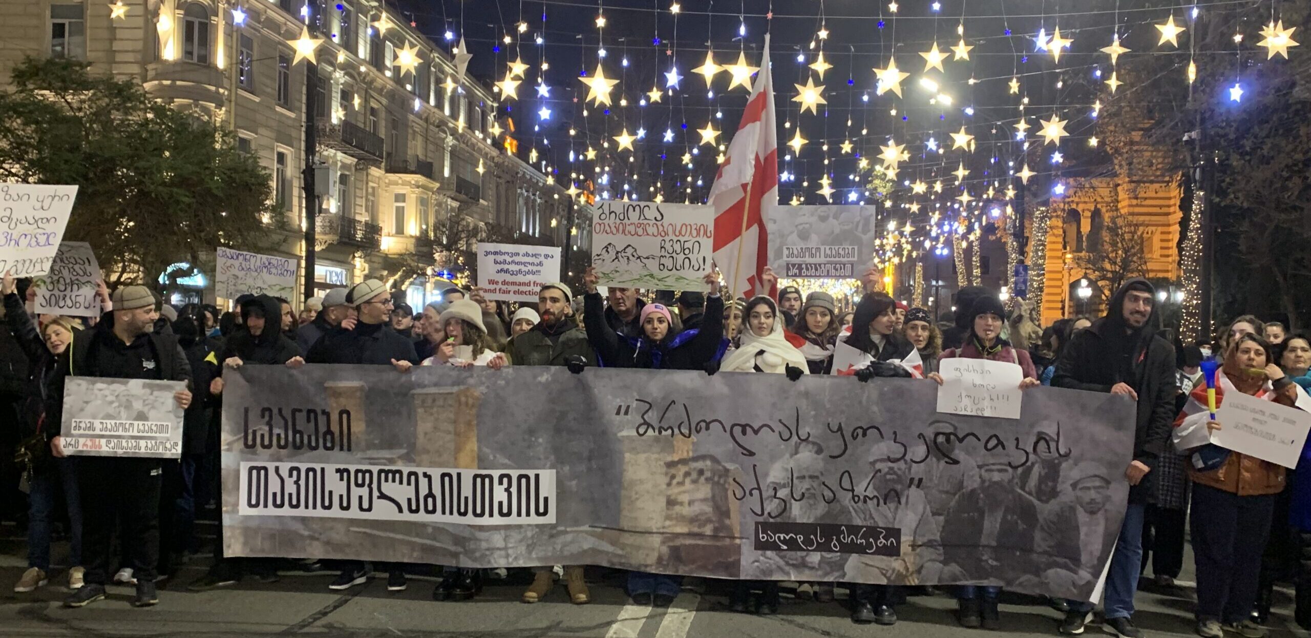 Menschen stehen mit einem Transparent auf der Straße. Auf einem Schild steht: Freie und Faire Wahlen!