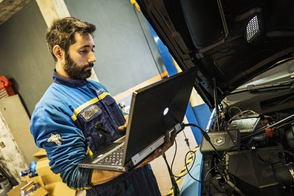 Mann mit Bart steht mit Laptop vor geöffneter Motorhaube