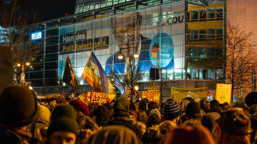 Menschen protestieren vor der CDU-Zentrale, in der ein riesiges Plakat von Friedrich Merz hängt.