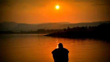 Eine Person in Silouette kniet vor einem Gewässer, das von einer tief hängenden Sonne orange erleuchtet wird.