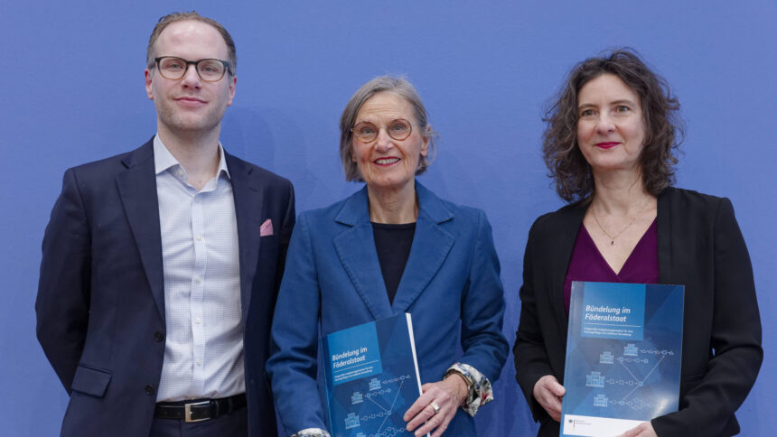 drei Menschen in förmlicher Kleidung stehen vor einer Pressewand, zwei halten ein Heft in der Hand