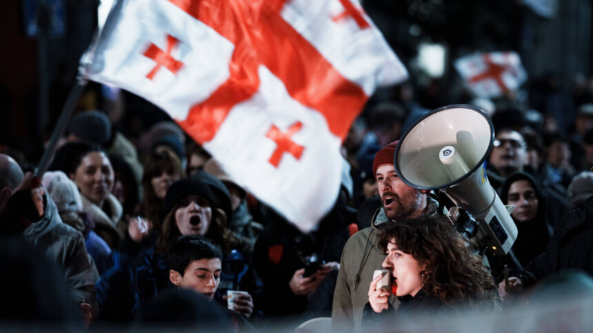 Frau hält Megafon in der Hand, im Hintergrund Menschen und eine große georgische Flagge.