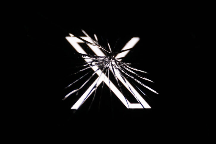 Ein Bildschirm mit vielen Sprüngen im Glas zeigt das Logo des Kurznachrichtendienstes X.