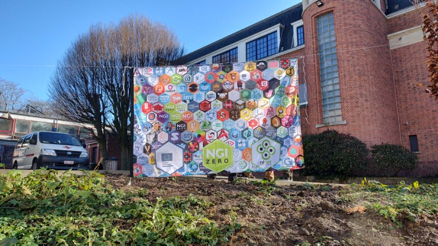 NGI Zero-Banner vor einem Van des Roten Kreuzes und einem roten Gebäude. Das Banner besteht aus vielen Sechsecken mit verschiedenen Logos.