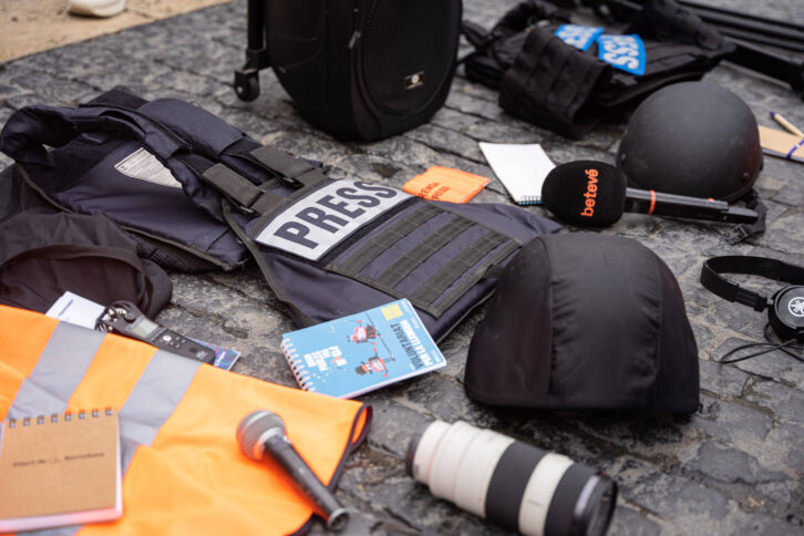 Ein Bild von einer Demonstration in Barceolna 2023, um Journalist:innen zu gedenken, die durch die israelische Armee getötet wurden. Auf der Erde liegen diverse Werkzeuge von Medienschaffenden wie Objektive und Mikrofone, in der Mitte eine blaue Schutzwese mit dem Wort "Press"
