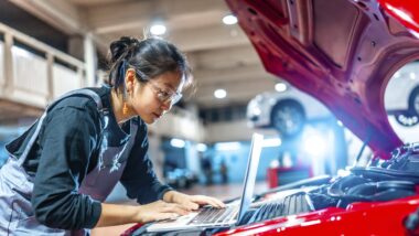 Junge Mechanikerin, die einen Laptop benutzt, während sie einen Automotor in einer professionellen Autowerkstatt überprüft.