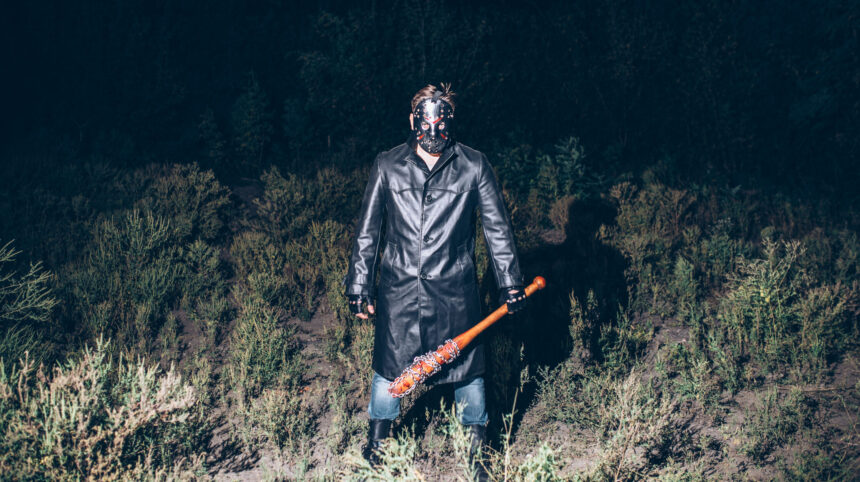 Ein Mensch mit Maske, Ledermantel und Baseballschläger, scharf angeblitzt auf einem nächtlichen Feld.