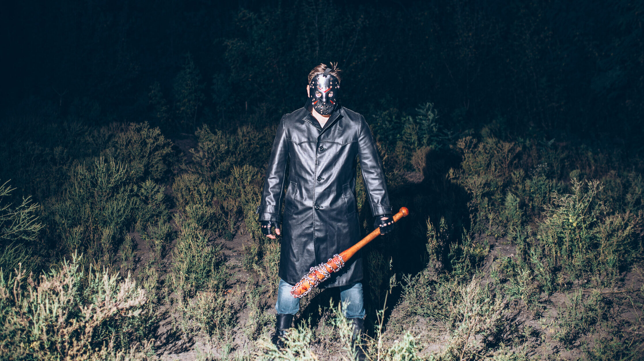 Ein Mensch mit Maske, Ledermantel und Baseballschläger, scharf angeblitzt auf einem nächtlichen Feld.