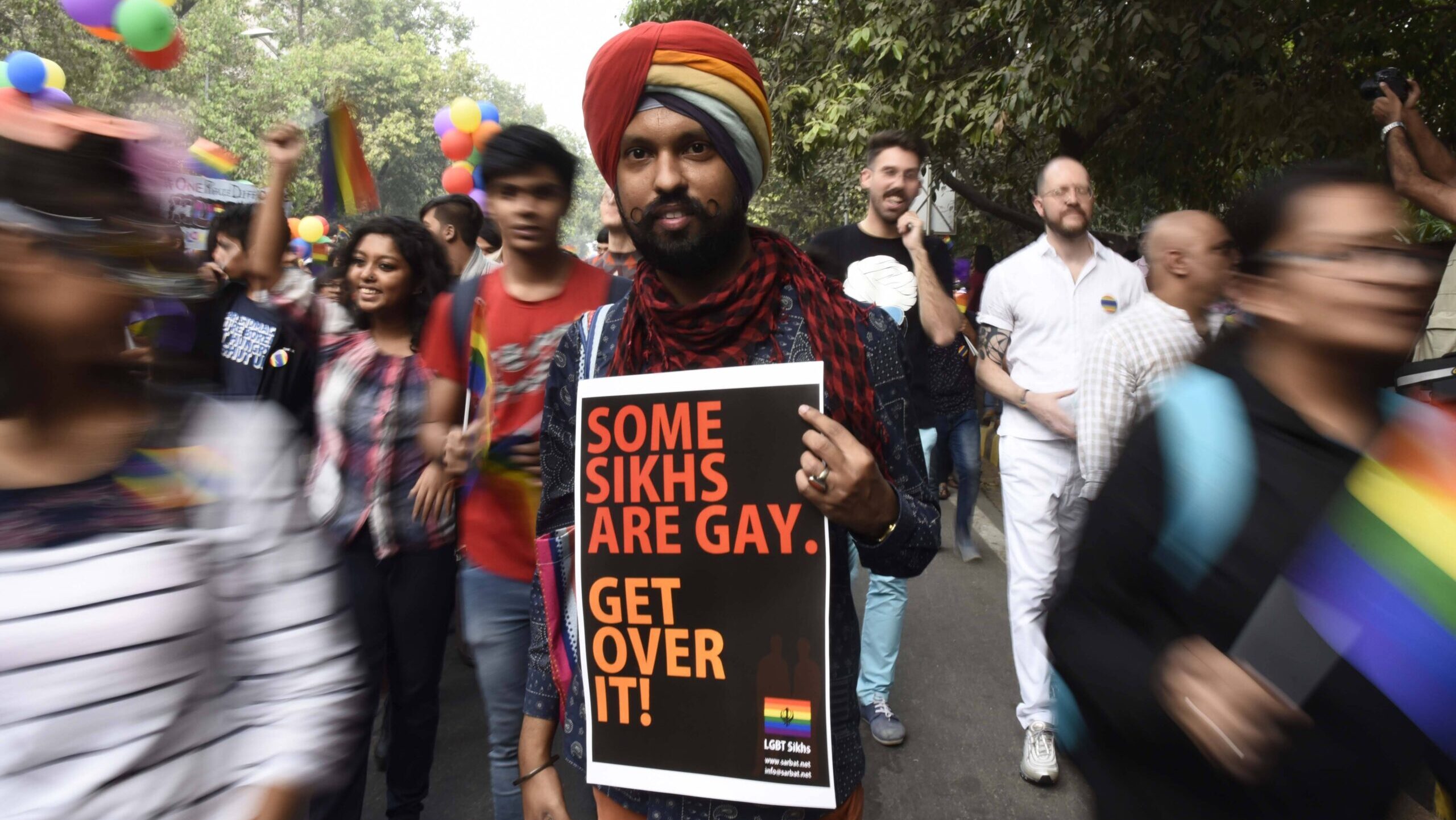 Fotografie eines Mannes mit Turban auf einer Queer Pride Demonstration. Der Mann zeigt ein Schild mit dem Text "Some Sikhs are gay. Get over it!". Im Hintergrund sind weitere bunt gekleidete Menschen, die zum Teil farbige Luftballons tragen.