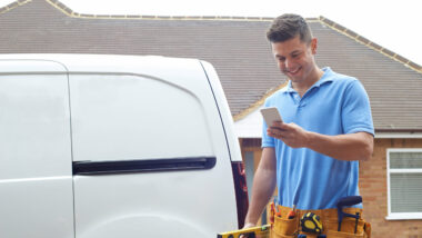 Ein Handwerker steht neben einem Transport-Fahrzeug. Er schaut auf sein Smartphone und lacht.