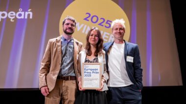 Drei Menschen lächeln auf einer Bühne und halten eine Urkunde, im Hintergrund erkennt man den Schriftzug "025 / Winner"