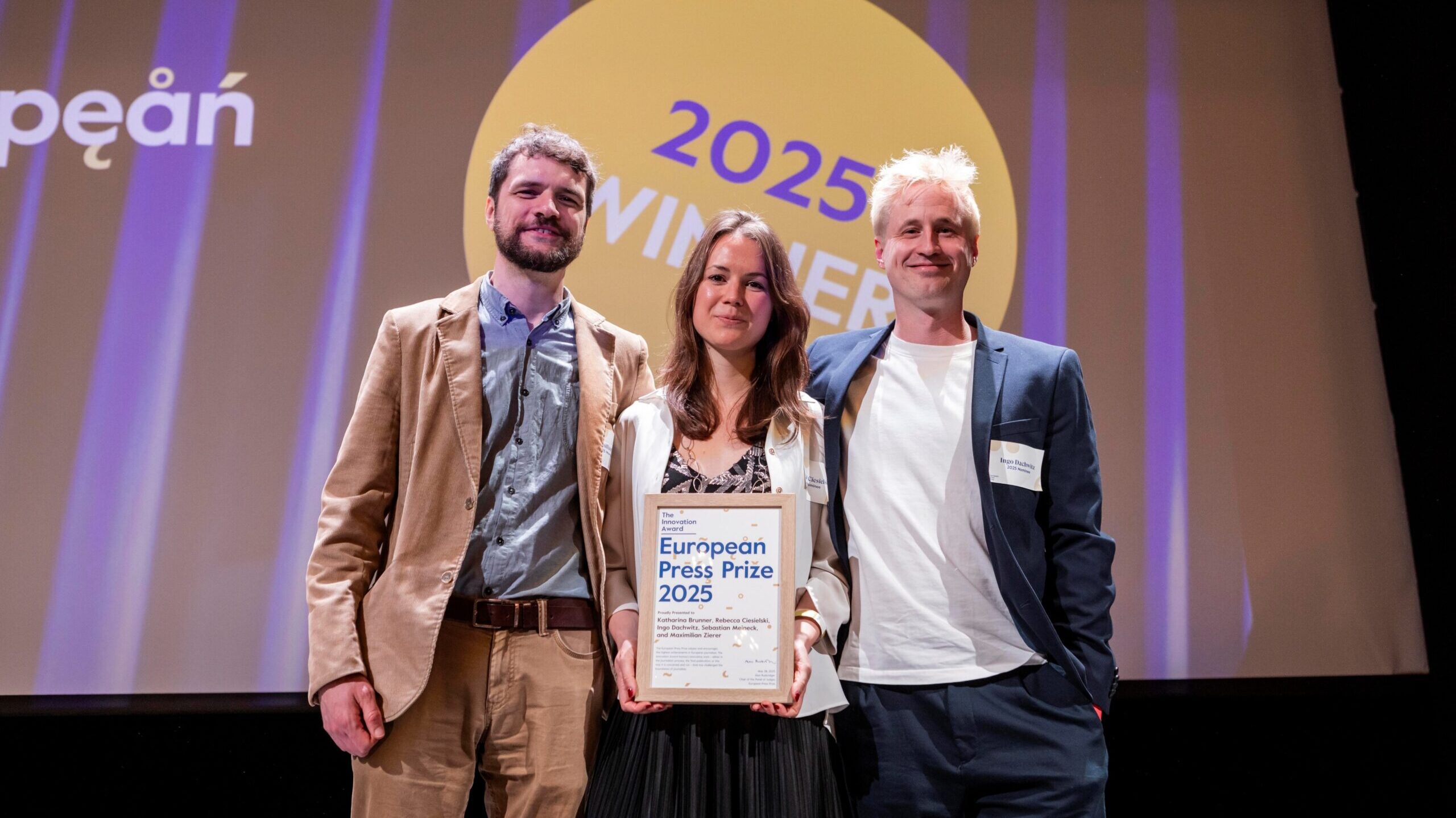 Drei Menschen lächeln auf einer Bühne und halten eine Urkunde, im Hintergrund erkennt man den Schriftzug "025 / Winner"