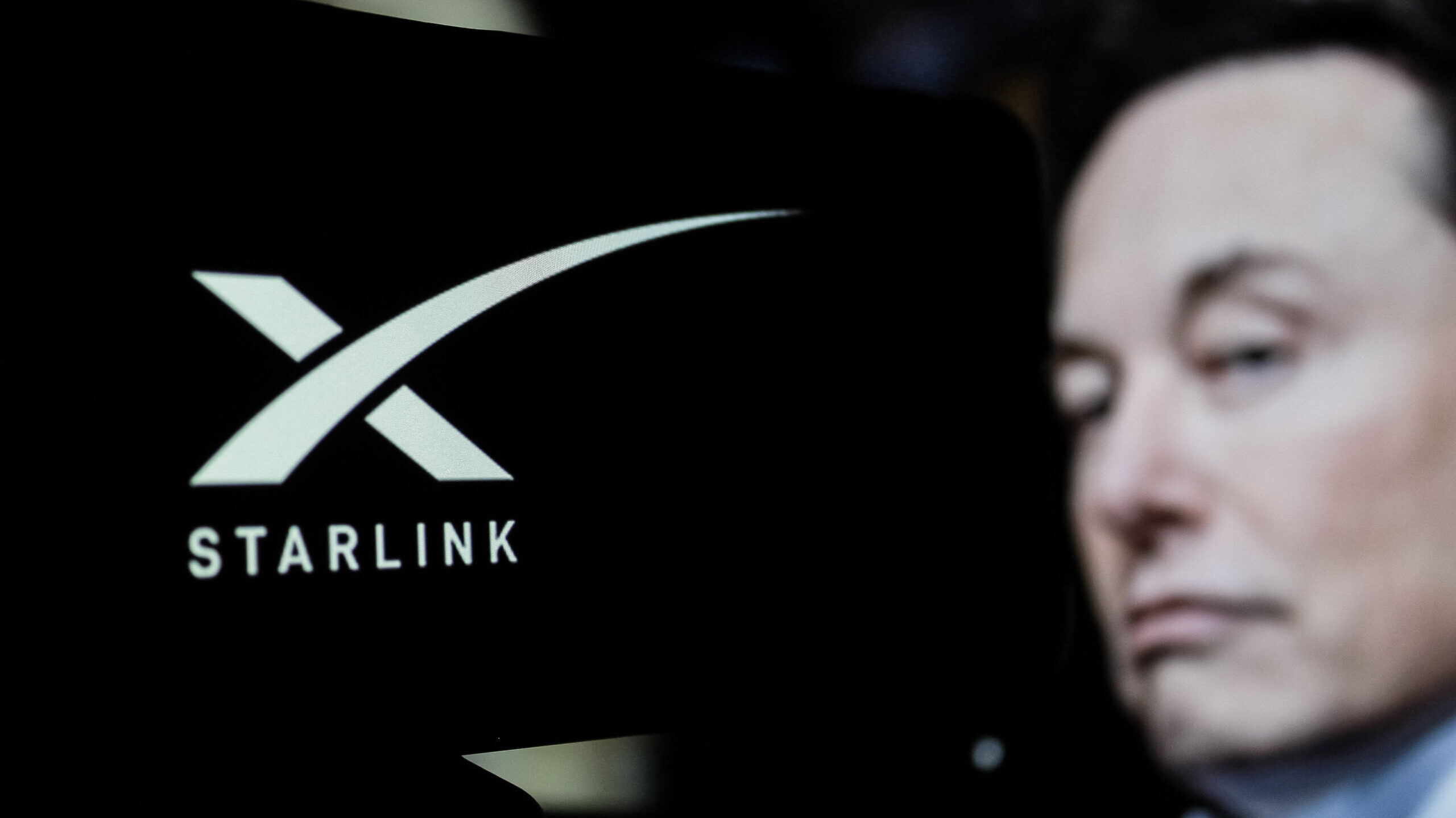 Ein unvorteilhaftes Foto eines blassen Elon Musk vor dem Starlink-Logo mit einem großen X