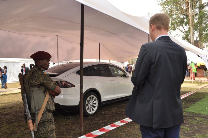 Ein weißes Luxusauto von Tesla unter einer weißen Plane. Davor stehen ein Geschäftsmann und ein Soldat, der das Auto bewacht