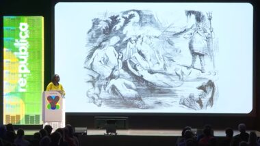 Eine Frau in gelbem Kleid auf einer großen Bühne, auf einer großen Leinwand ist eine Zeichnung zu sehen, die Neptun und Meerjungfrauen mit einem Unterseekabel darstellt. Links von der Bühne steht "re:publica"