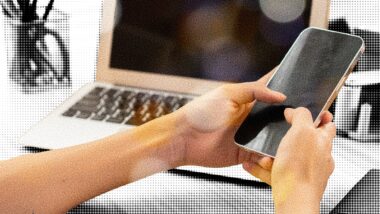Eine Person hält ein Smartphione, dahinter ist ein Laptop zu sehen.
