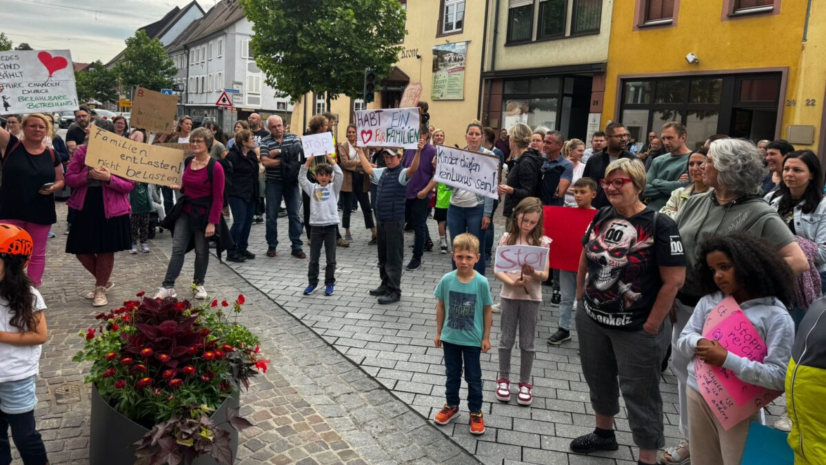 Familien mit Schildern demonstrieren gegen hohe Kosten für Kindergarten.
