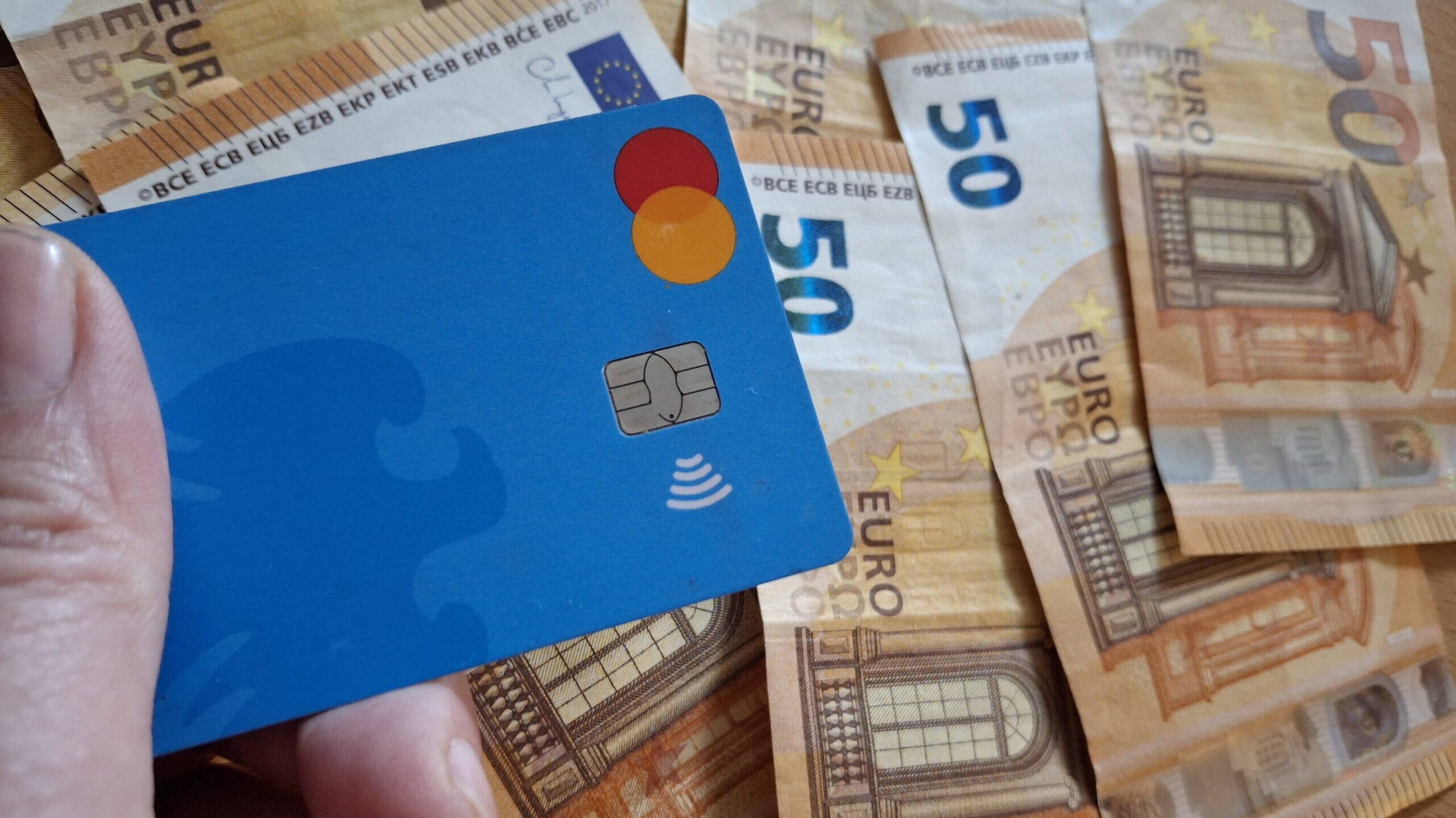 Eine Hand hält eine Bankkarte, dahinter liegt Bargeld.
