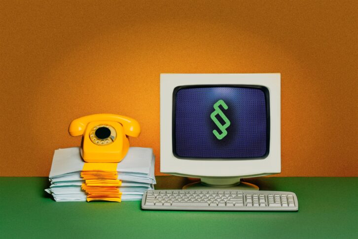 Schreibtisch, auf dem ein Röhrenbildschirm mit einem Paragraf-Zeichen, davor eine Tastatur; daneben ein Stapel Papier, auf dem ein Telefon mit Wählscheibe steht