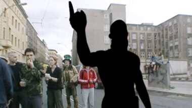 Das Bild zeigt eine Straßenszene in Berlin, an einem bedeckten, aber hellen Tag. Im Hintergrund sind mehrere mehrstöckige Gebäude zu sehen, die einen etwas heruntergekommenen Eindruck machen. Die Gebäude sind hellbeige und zeigen den Zahn der Zeit. Im Vordergrund steht eine Person, deren Gesicht und Oberkörper durch eine schwarze Silhouette verdeckt sind. Nur die Arme und Hände sind zu sehen. Die Person hebt den rechten Arm und zeigt mit dem Zeigefinger nach oben, was auf eine Geste von Führung, Protest oder Ankündigung hindeutet. Die Silhouette suggeriert eine gewisse Macht oder Autorität, gleichzeitig aber auch Anonymität und Geheimnis. Vor der Person steht eine kleine Gruppe von etwa sieben bis acht Menschen, die unterschiedlich gekleidet sind. Sie scheinen sich zu unterhalten oder zu warten. Es gibt Männer und Frauen, einige tragen Alltagskleidung, andere erscheinen eher in Freizeitkleidung. Ein Mann trägt einen grünen Hut, der an einen Leprechaun-Hut erinnert. Die Personen schauen in verschiedene Richtungen, wobei einige direkt auf die silhouettierte Person blicken, andere nicht. Zwei junge Frauen sitzen auf einer niedrigen Mauer oder einem Betonblock am rechten Bildrand. Die Mauer ist etwas verwittert und zeigt Graffiti-Spuren. Die allgemeine Atmosphäre ist eher ruhig und neutral, obwohl die Geste der silhouettierten Person eine gewisse Spannung oder Dynamik in das Bild bringt. Die Stimmung ist etwas unklar, sie könnte als kritisch, nachdenklich oder auch einfach als dokumentarisch interpretiert werden.