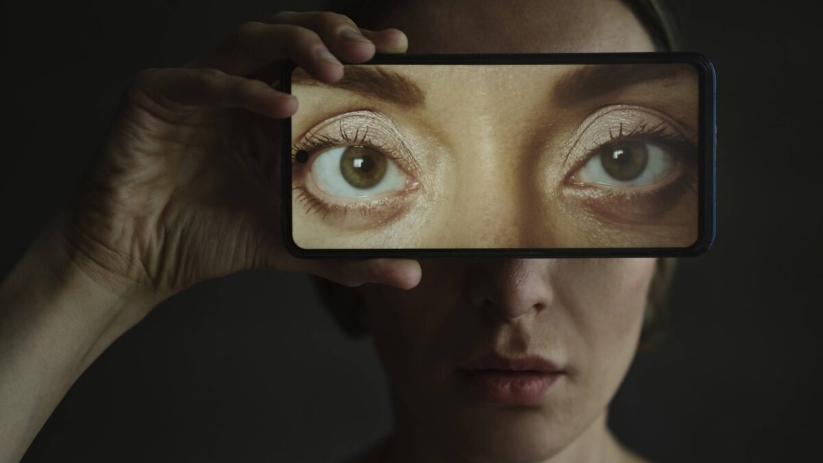 Eine Person hält sich ein Smartphone vors Gesicht, dessen Display ein Augenpaar zeigt.