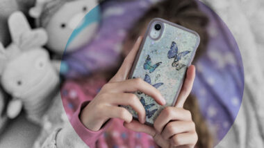 Jugendilche Person liegt im Bett und hält ein Handy vors Gesicht. Die Rückseite des Handys ist mit Schmetterlingen dekoriert.