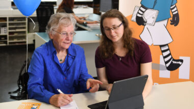 Eine ältere und eine jüngere Frau sitzen vor einem Tablet mit Tastatur. Die ältere zeigt auf etwas und macht sich mit der anderen Hand eine Notiz mit einem Stift auf einem kleinen Block.