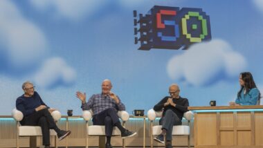 Drei Männer sitzen auf einer Bühne vor himmelblauem Hintergrund, auf dem eine große bunte Zahl 50 prangt.