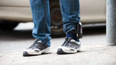 Beine in Jeans und Turnschuhen, an einem Knöchel ist eine elektronische Fußfessel befestigt