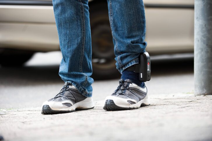 Beine in Jeans und Turnschuhen, an einem Knöchel ist eine elektronische Fußfessel befestigt