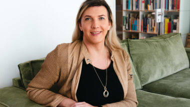 Julia Monro sitzt auf einem grünen Sofa und lächelt leicht in die Kamera. Im Hintergrund sind Bücherregale.